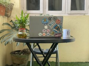 Laptop adorned with vibrant stickers illustrating a WordPress enthusiast's journey, on a table with a lucky bamboo plant and a mug on each side. Each sticker tells a story of achievements and community love.
